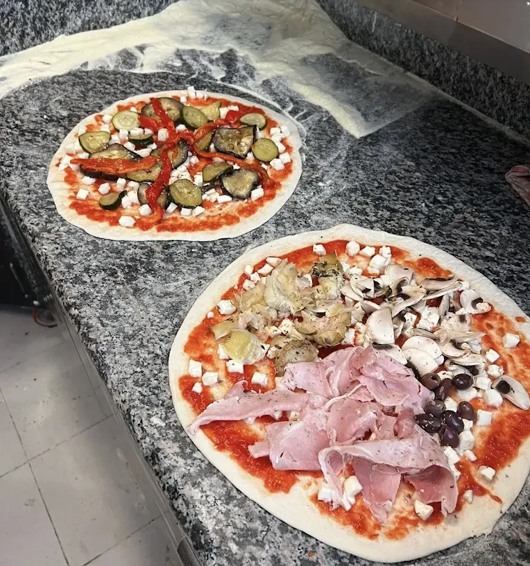 Two pizzas being topped with vegetables, ham, mushrooms and olives