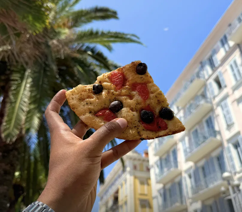 A slice of focaccia held up against palm trees and Nice buildings
